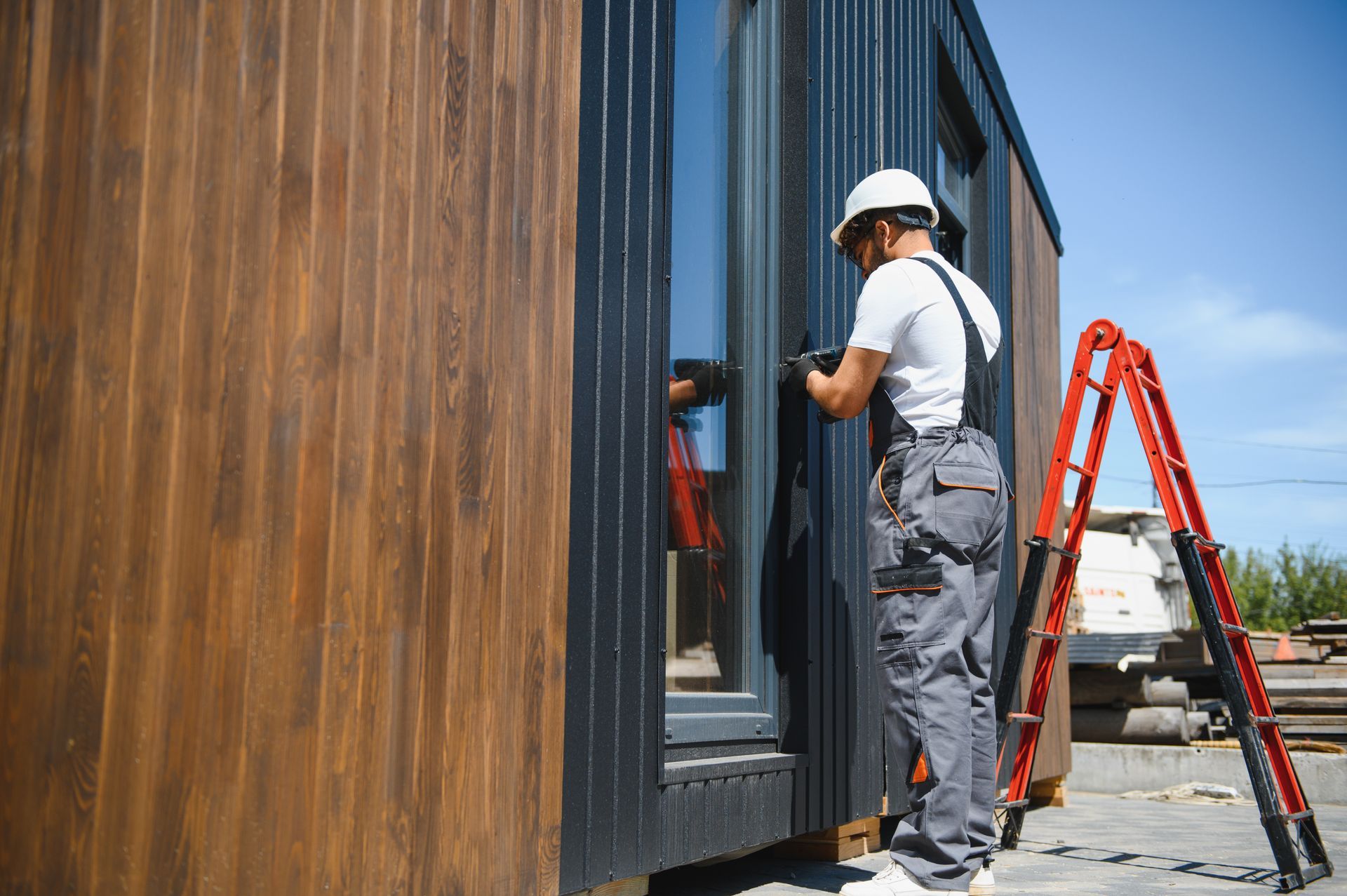 A construction worker in a hard hat and grey overalls works on the side of a modern wooden building near a red ladder.