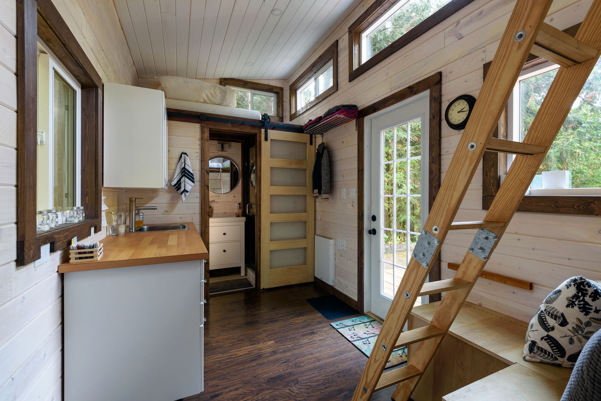 Interior of a tiny house with light wood walls, a ladder to a loft, a small kitchen counter, and a bathroom doorway.