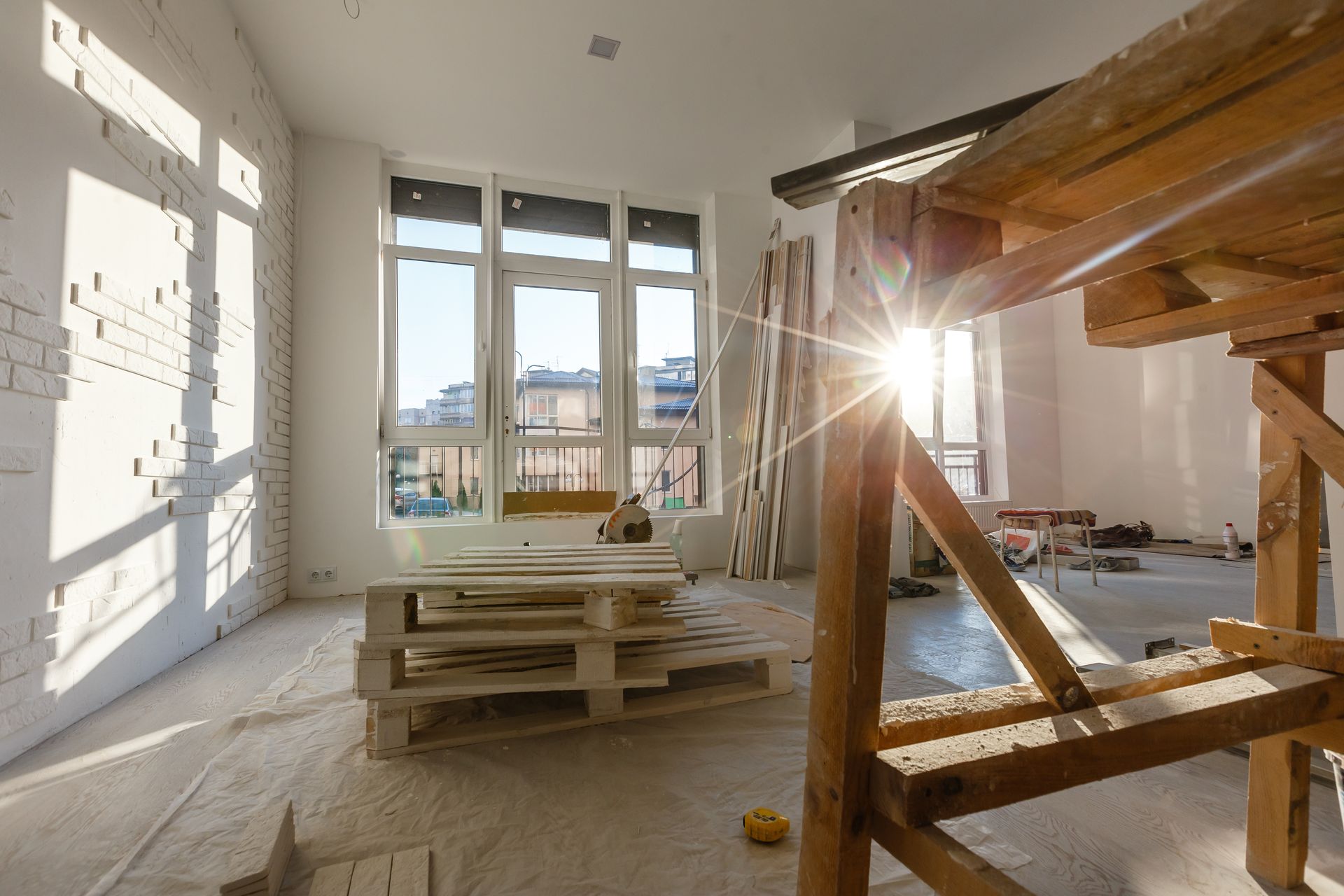 Sunlight streams through large windows into an unfinished room with white brick walls, wooden scaffolding, and pallets.