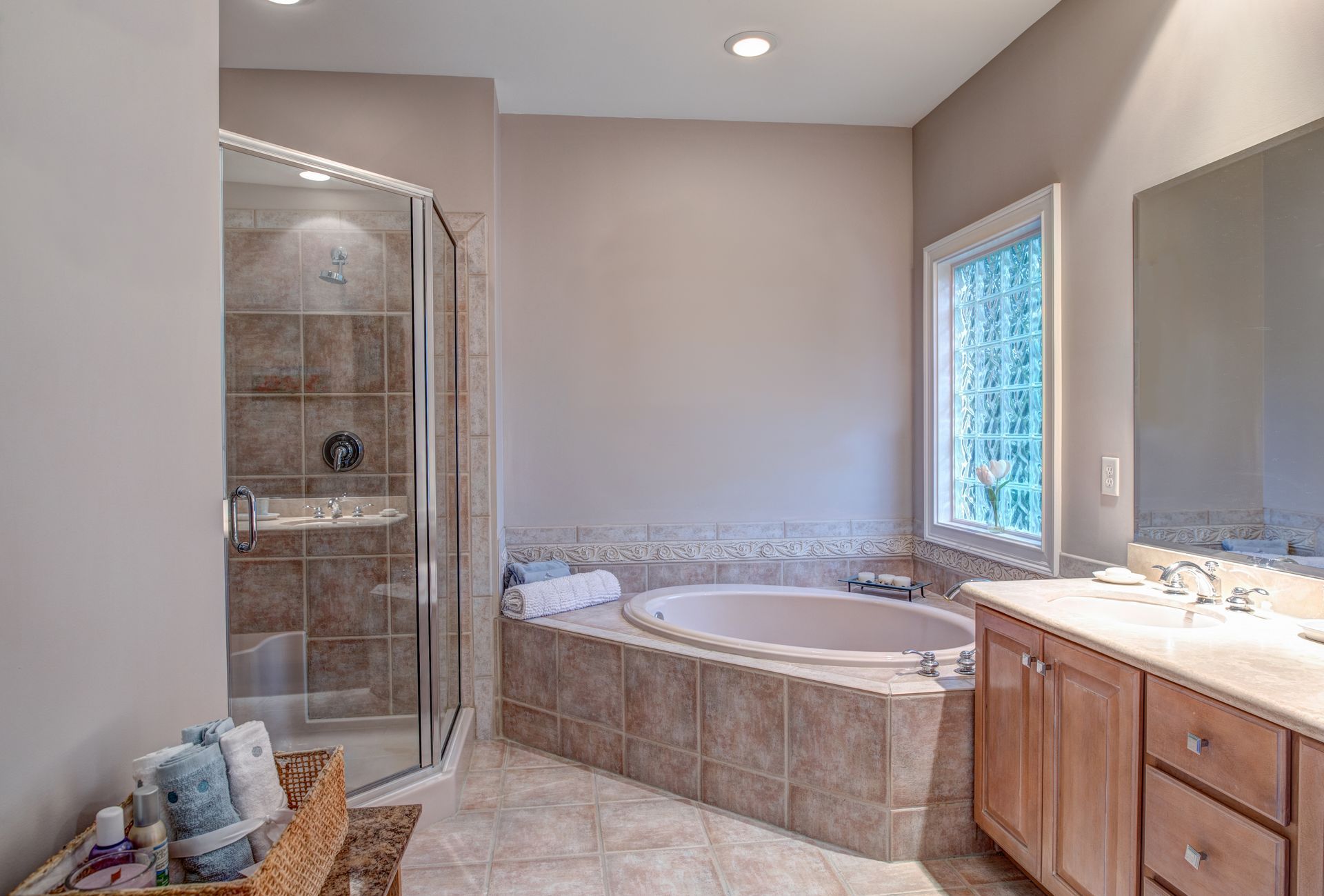 A beige bathroom featuring a tiled walk-in shower, a corner soaking tub, a wooden vanity with a mirror, and a window.
