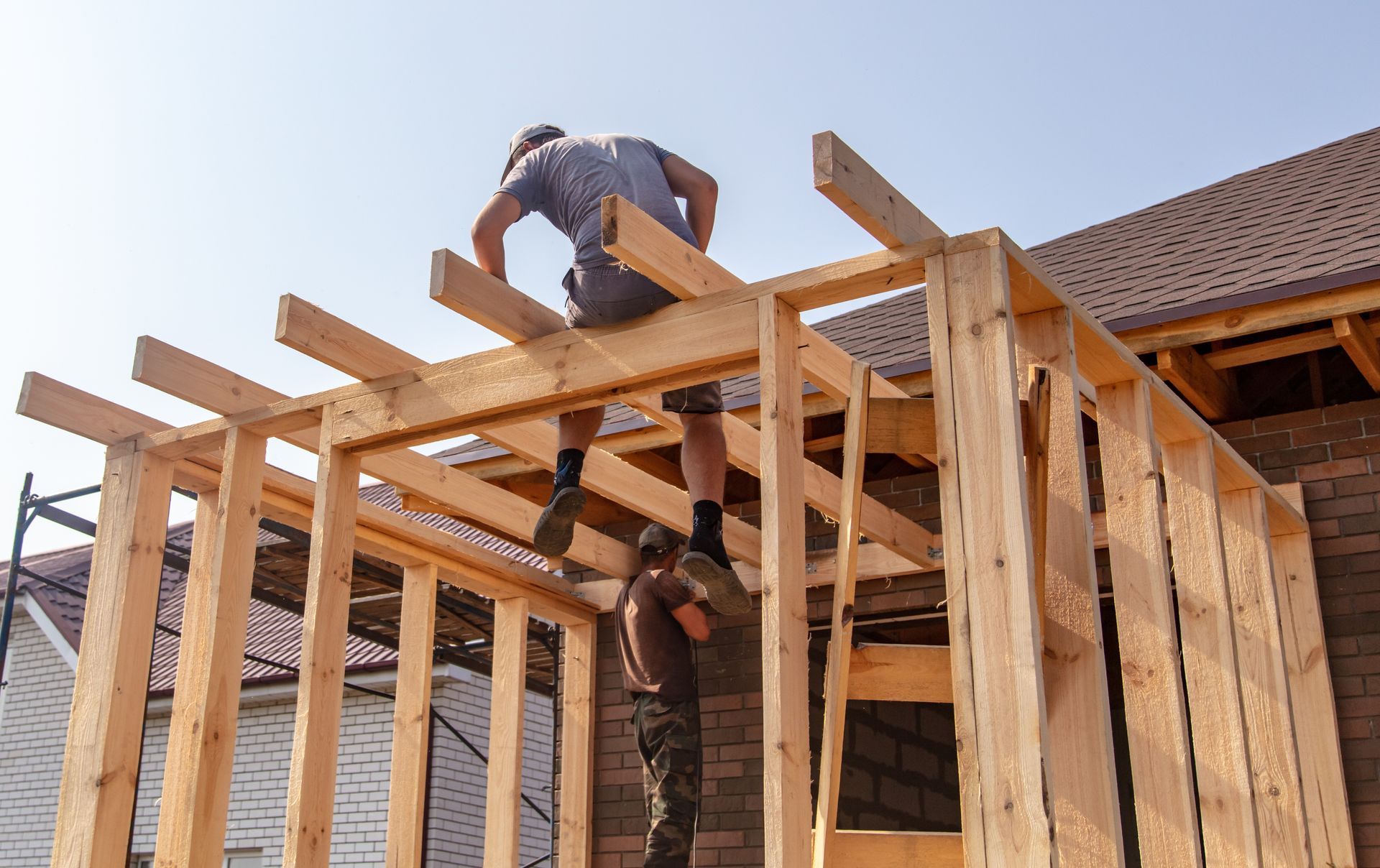 Two people construct the wooden frame of a house addition attached to an existing brick building.