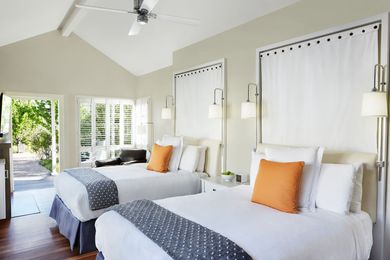 Two queen beds with white linens, orange accent pillows, and dark patterned runners in a bright, modern bedroom.