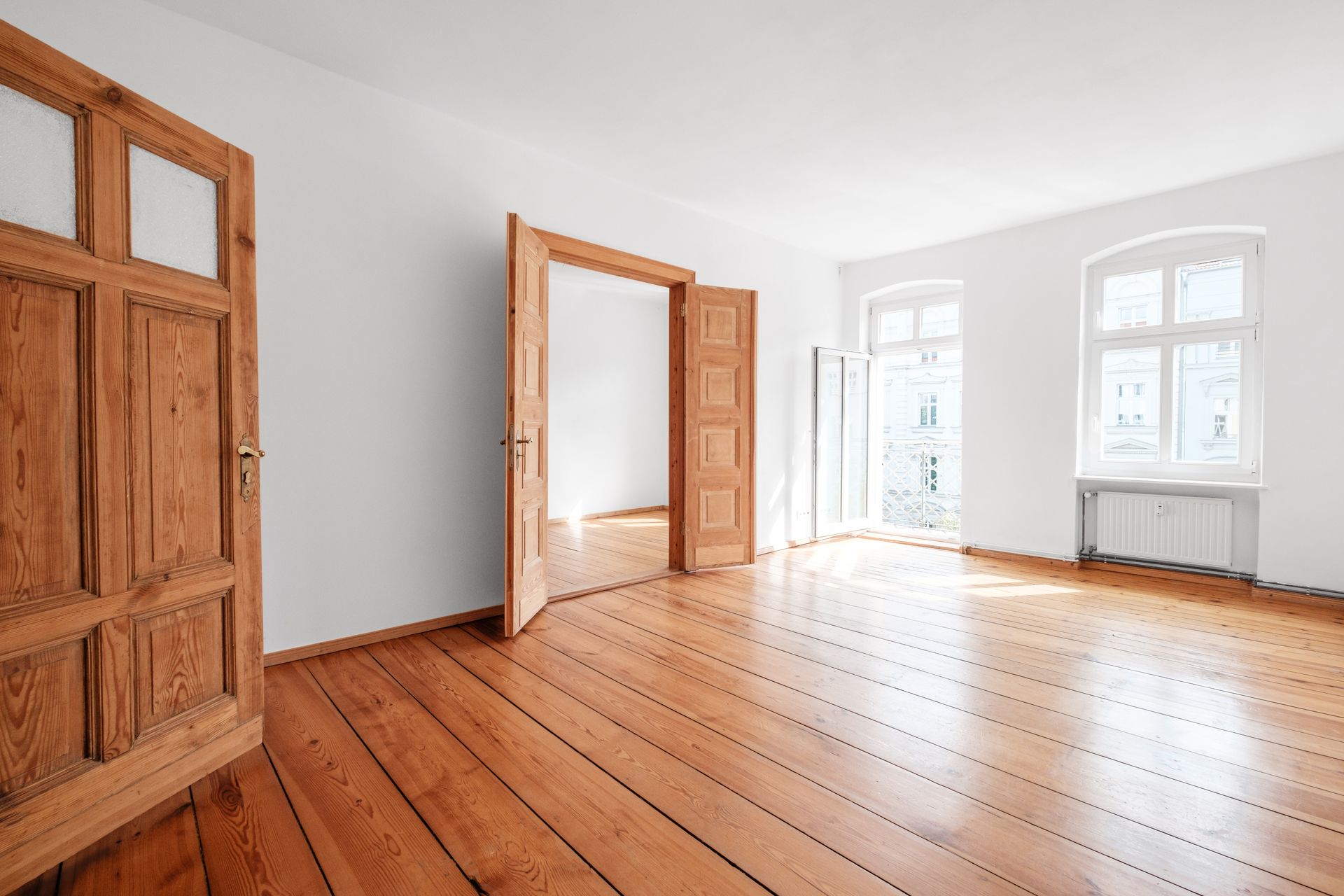 An empty room with light wood floors, white walls, large windows, and classic wooden panel doors.