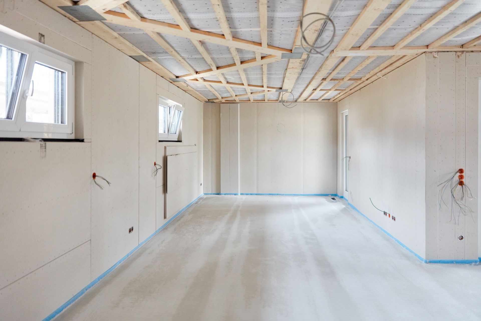 An interior room under construction with white plasterboard walls, a concrete floor, and exposed wooden ceiling beams.