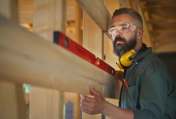 A carpenter uses a spirit level to check the alignment of a wooden beam while wearing safety glasses and ear protection.