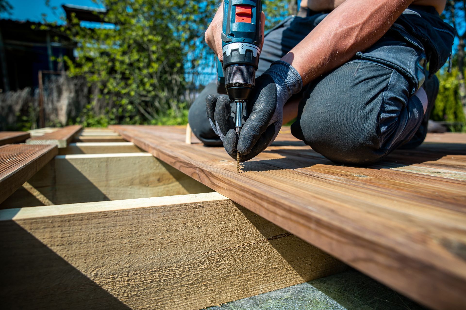 A person wearing gloves uses a cordless drill to fasten wooden planks onto a deck frame outdoors in sunlight.