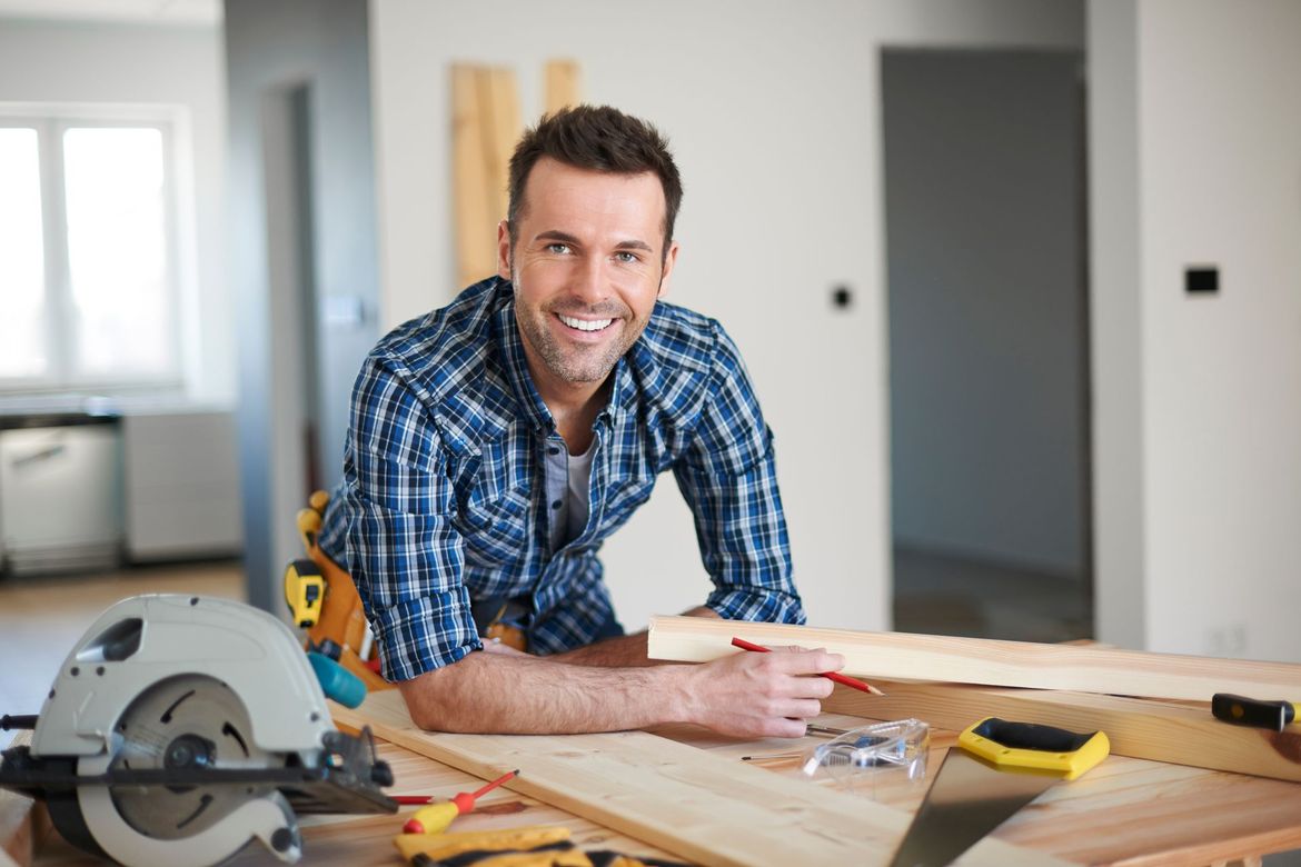 A smiling carpenter in a plaid shirt prepares to mark a piece of wood on a workbench with tools nearby.