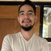 Man with dark hair and beard smiles, wearing a white shirt; set outside near a door.