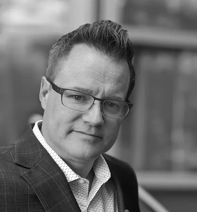 Man in a suit jacket and glasses, looking at the camera. He is in front of a blurry background.