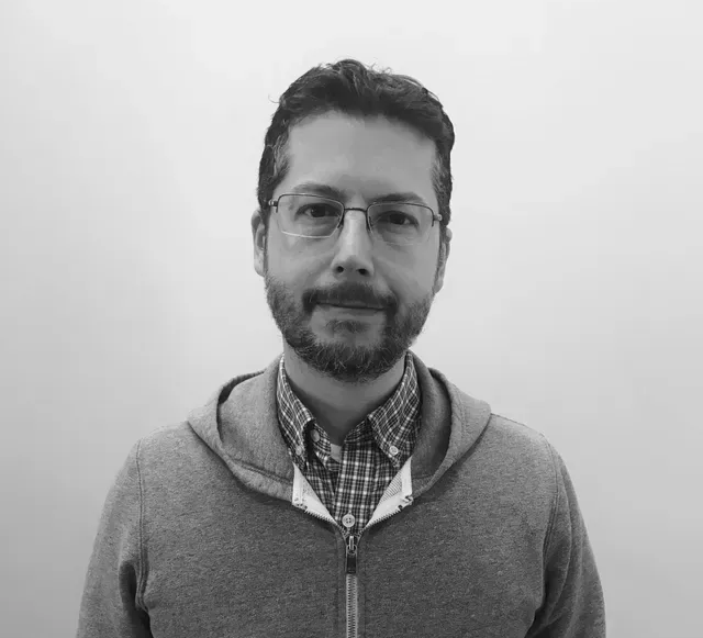 Man with glasses and beard, wearing a gray hooded sweatshirt over a plaid shirt, standing in front of a white wall.