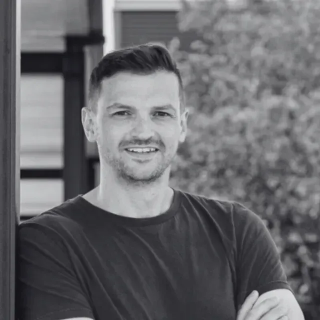 Man smiling, arms crossed, leaning on a pillar, wearing a dark shirt outdoors.