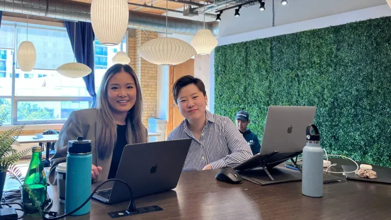 Two women are sitting at a table with laptops.