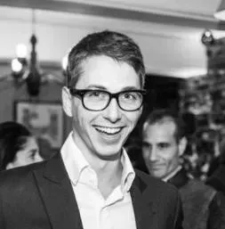 Man wearing glasses and a suit jacket, smiling, with another man in the background.