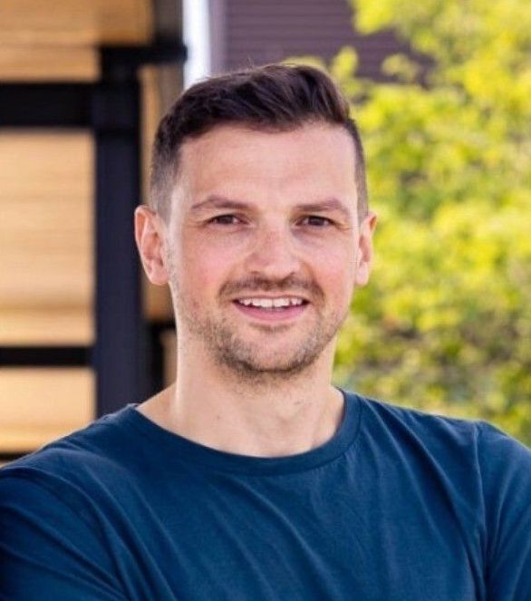 Man with short dark hair, smiling, wearing a blue t-shirt, outdoors near a wooden structure and foliage.