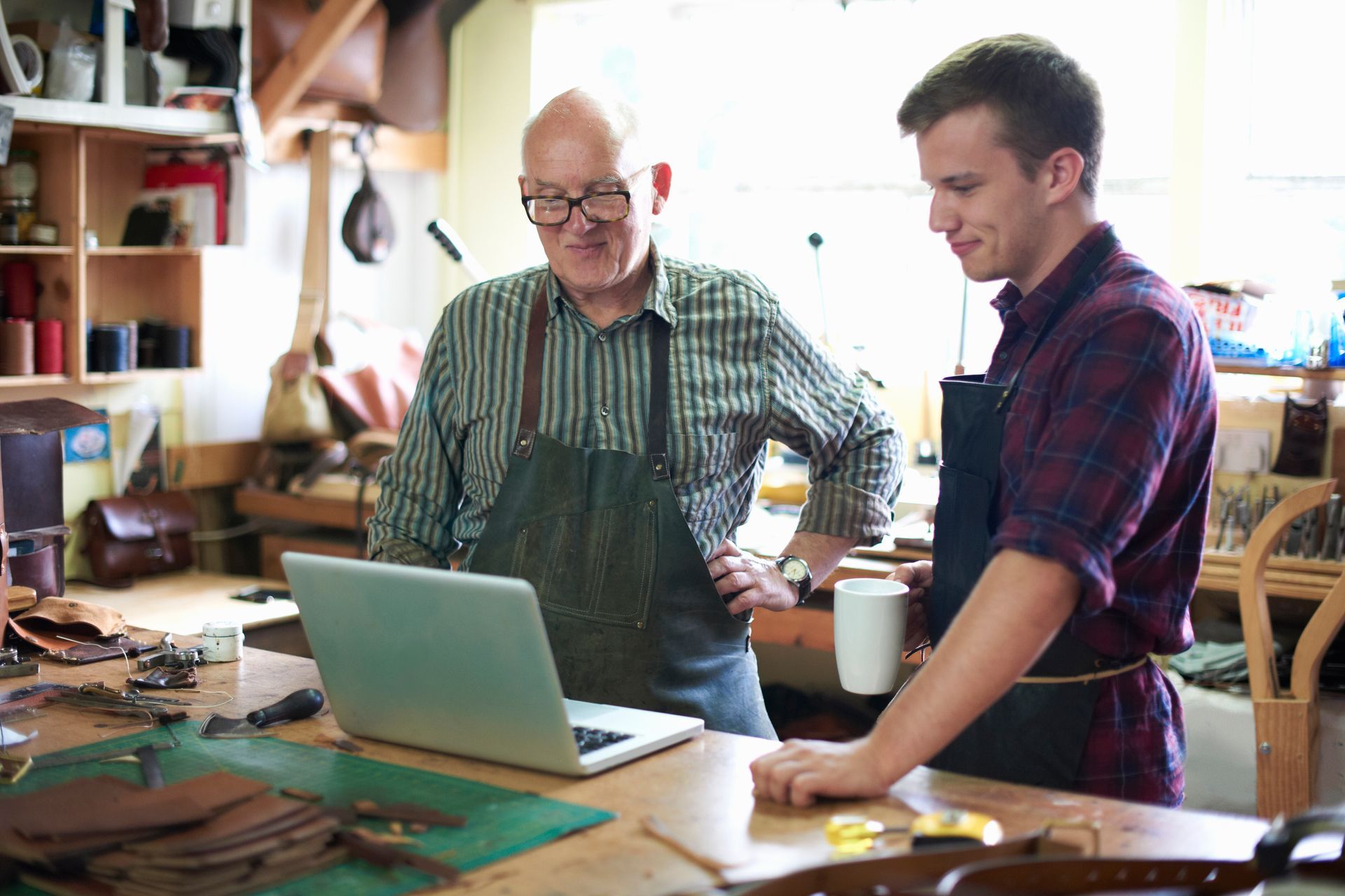 Two men looking at a laptop in a workshop; one older with glasses, the other younger with a coffee cup.