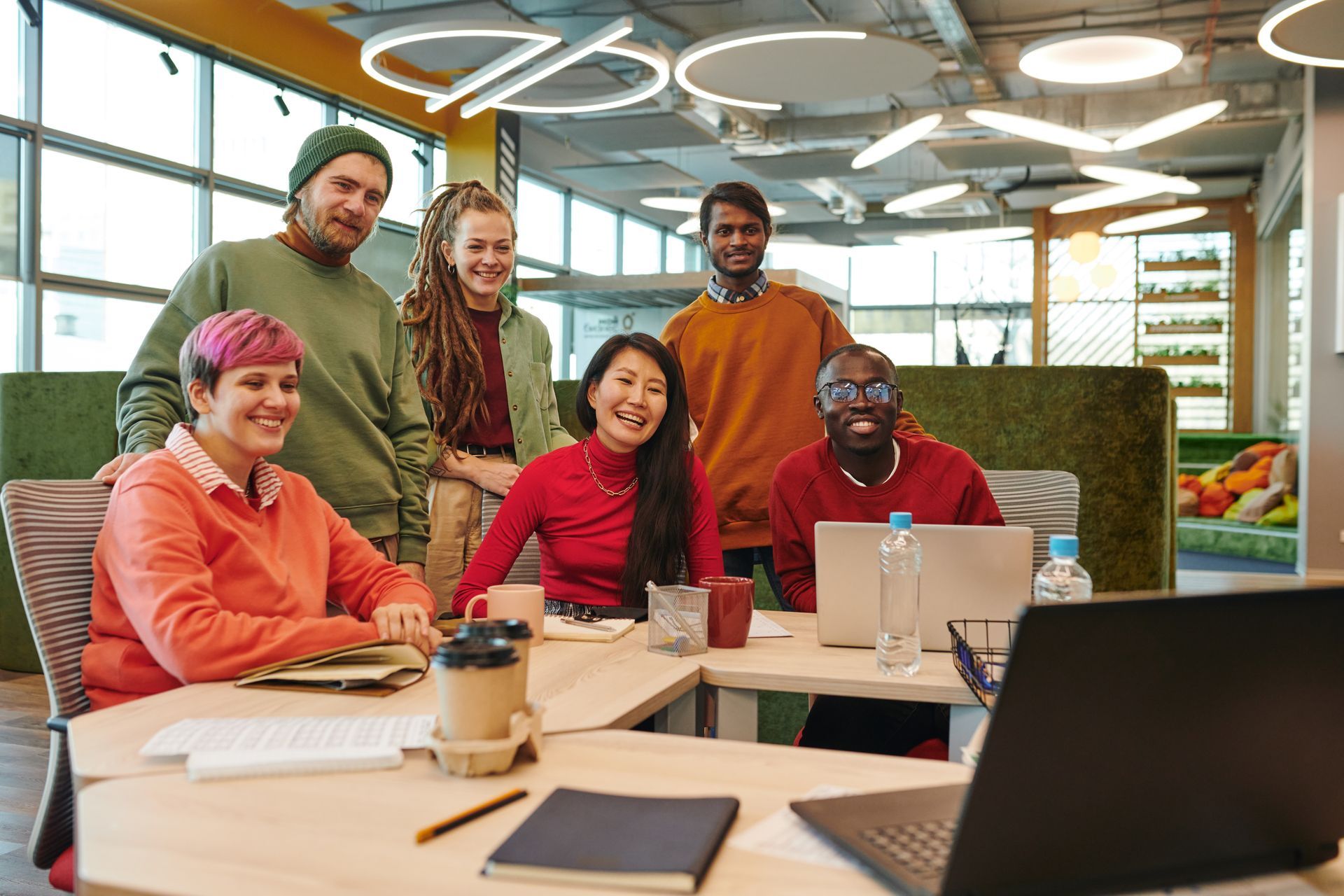 Group of people smiling at a table with laptops, coffee, and notebooks in a bright office setting.