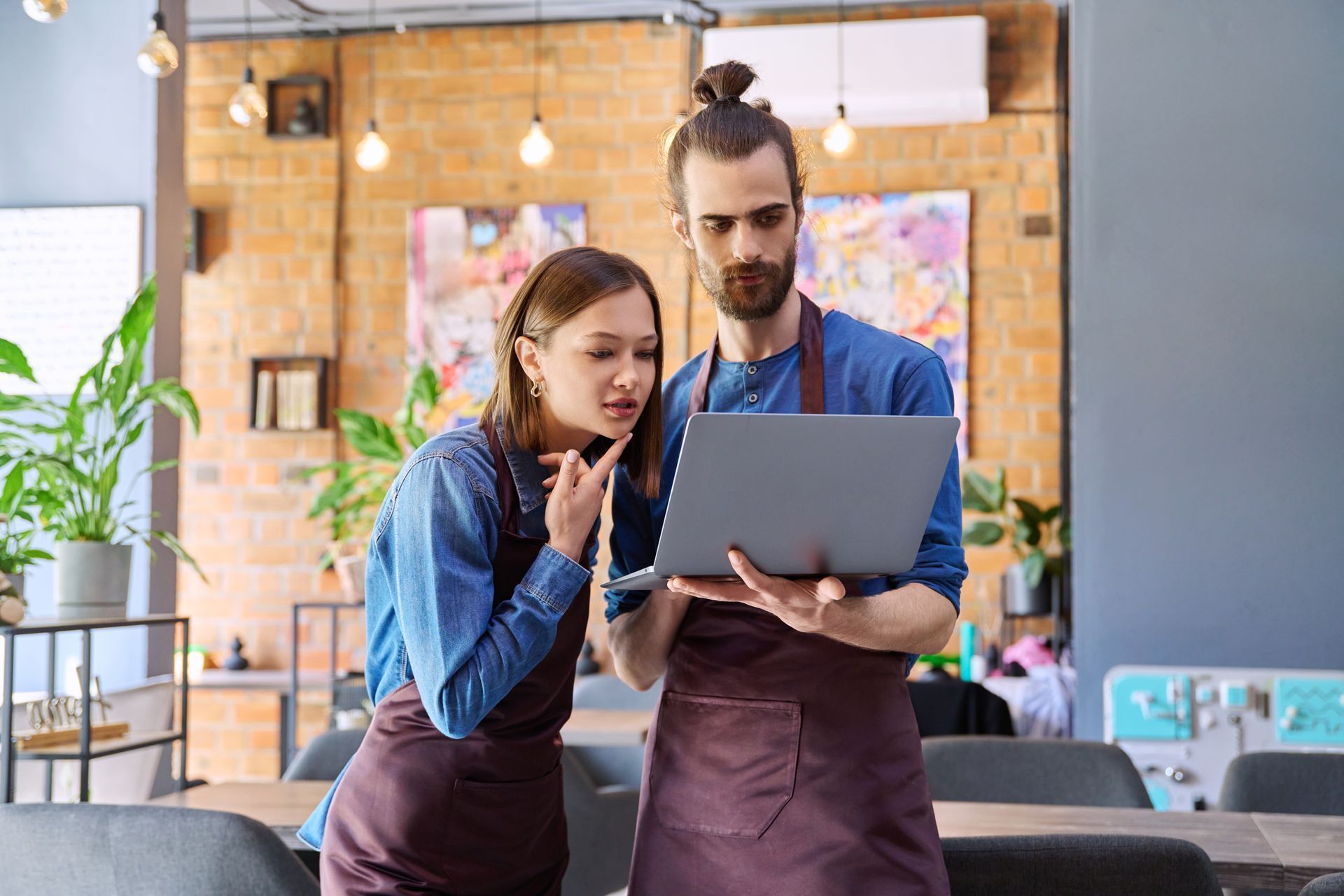 Two people in aprons look at a laptop in a cafe setting, with puzzled expressions.