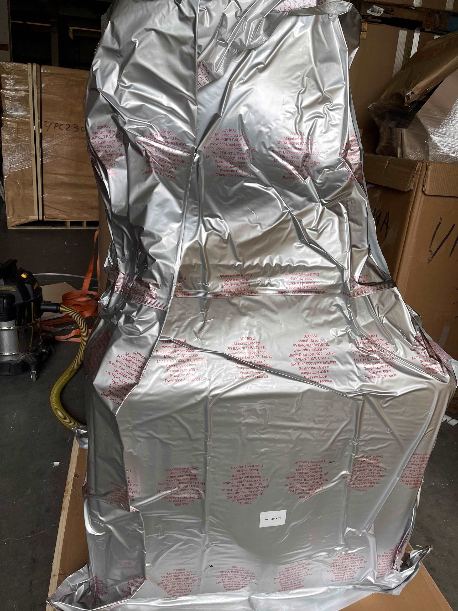 A large object wrapped in metallic, foil-like protective material, sitting on a wooden pallet in a warehouse setting.