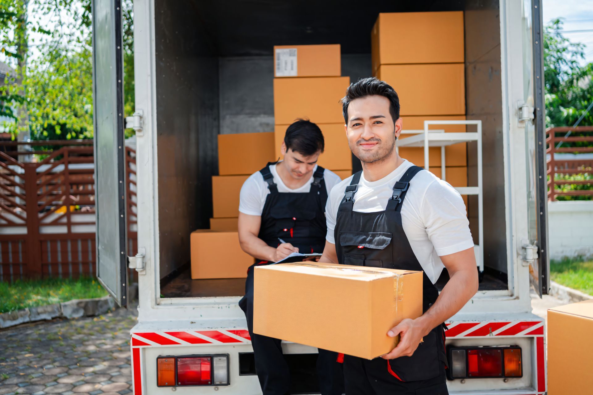 Two men are standing in front of a moving truck holding boxes.