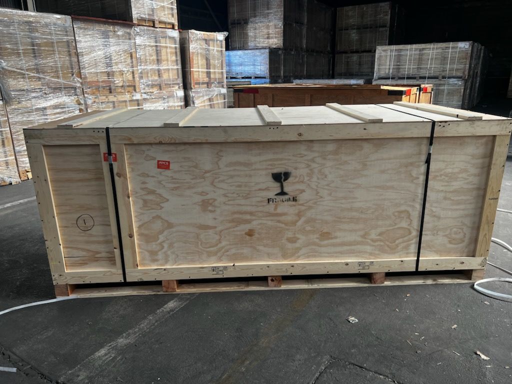 A large wooden shipping crate sits on a warehouse floor in front of stacked crates.