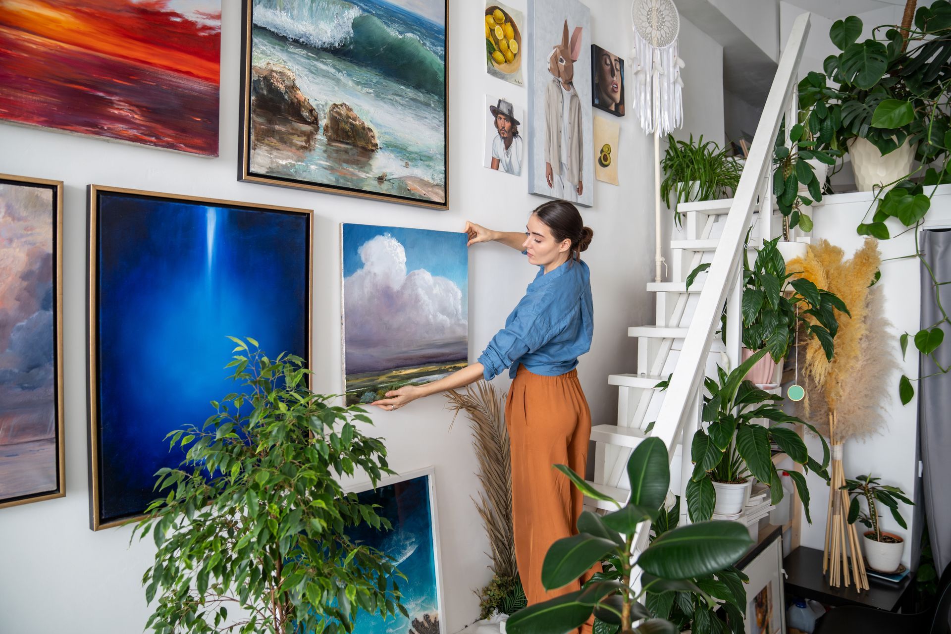 A woman is hanging a painting on a wall next to a staircase.
