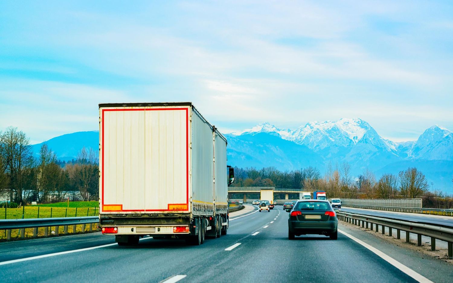 A white semi-truck and a dark car driving on a highway toward snow-capped mountains under a clear blue sky.