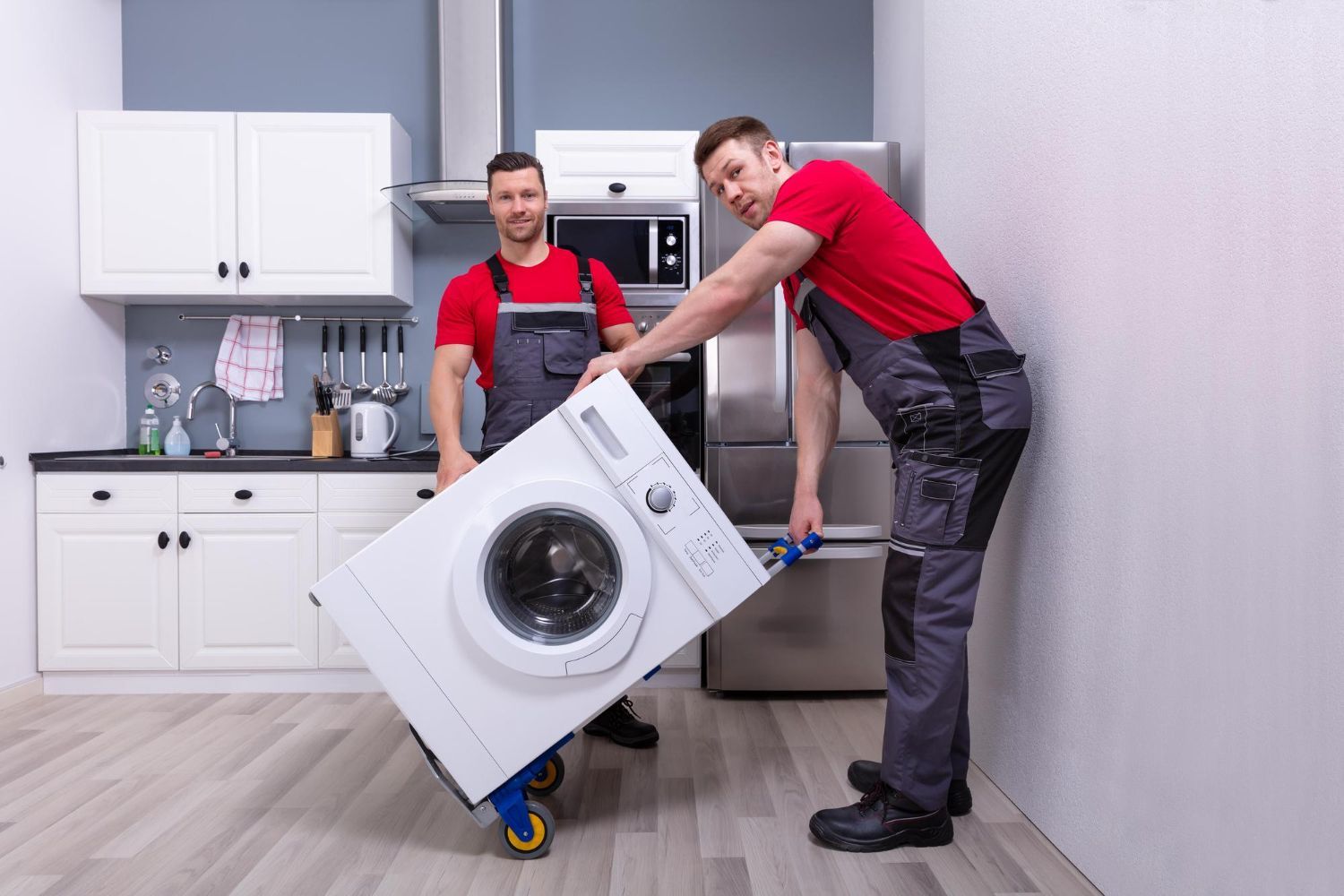 Two workers in red shirts and gray work overalls use a hand truck to move a white washing machine in a kitchen.