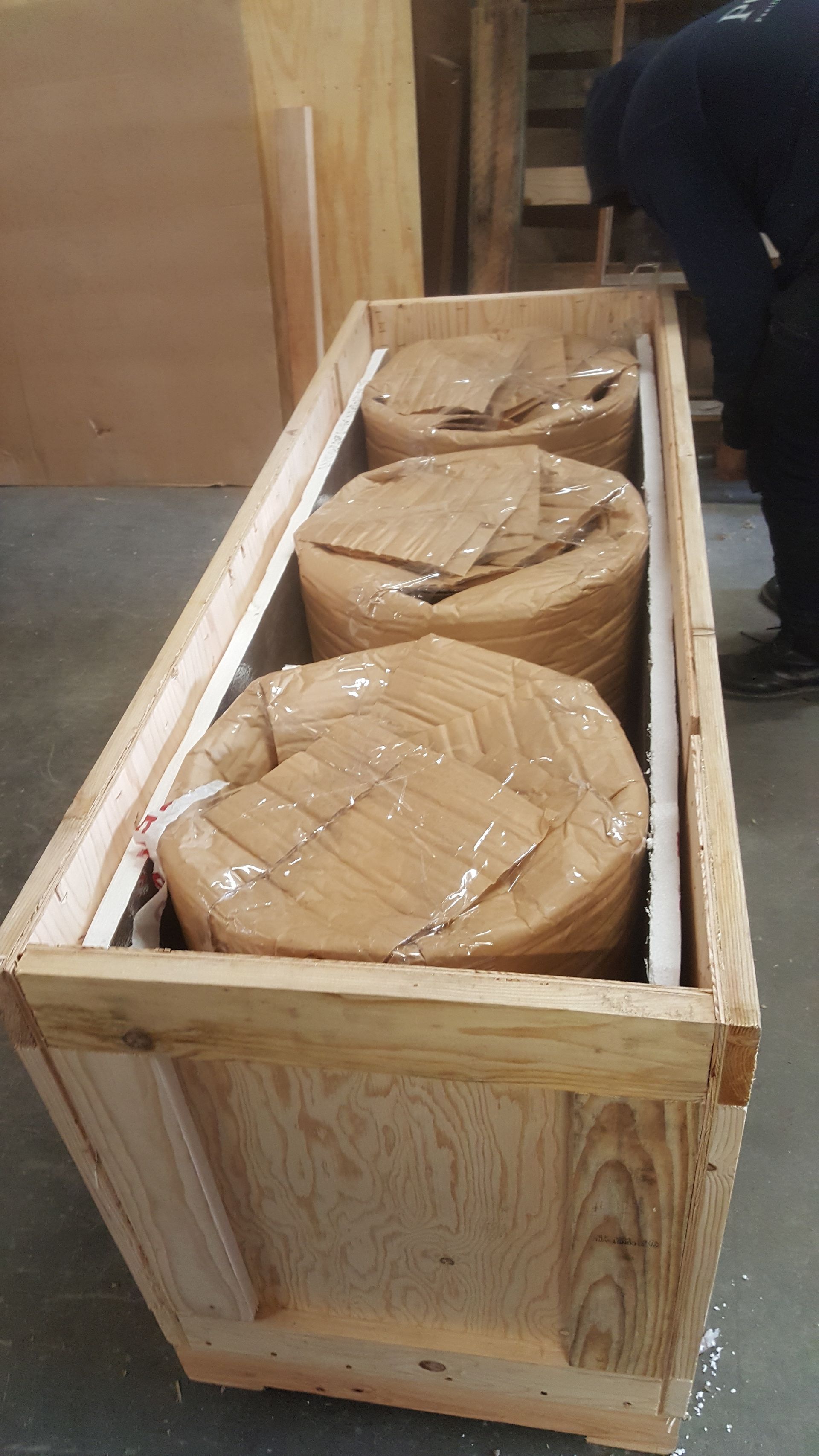 Three wrapped, cylindrical industrial items packed in a vertical wooden crate in a warehouse setting.