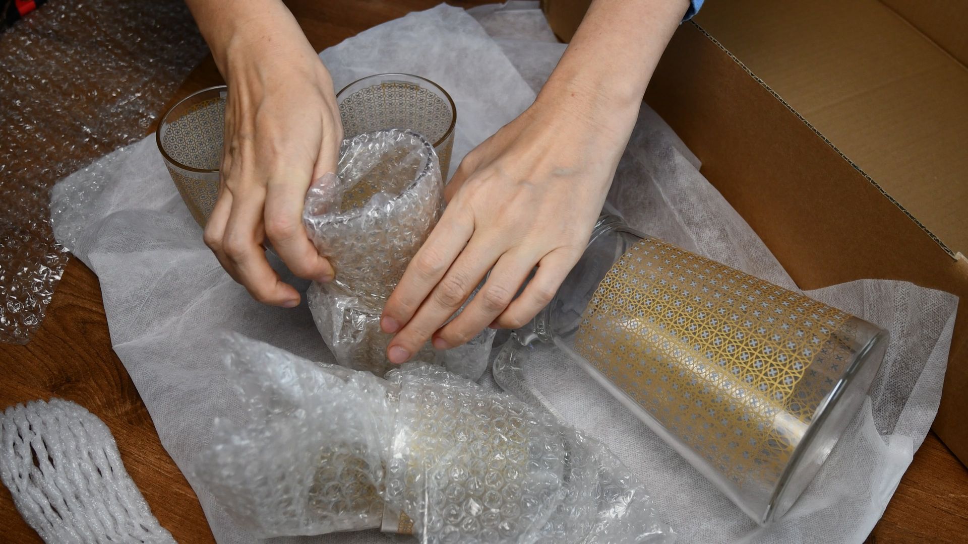 Hands wrapping glasses in bubble wrap inside a cardboard box, preparing for shipping.