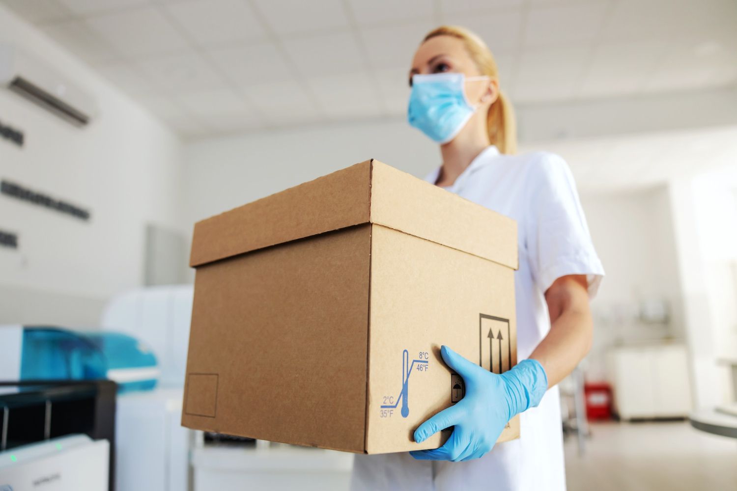 A healthcare worker wearing a mask and blue gloves holds a cardboard box in a clinic.