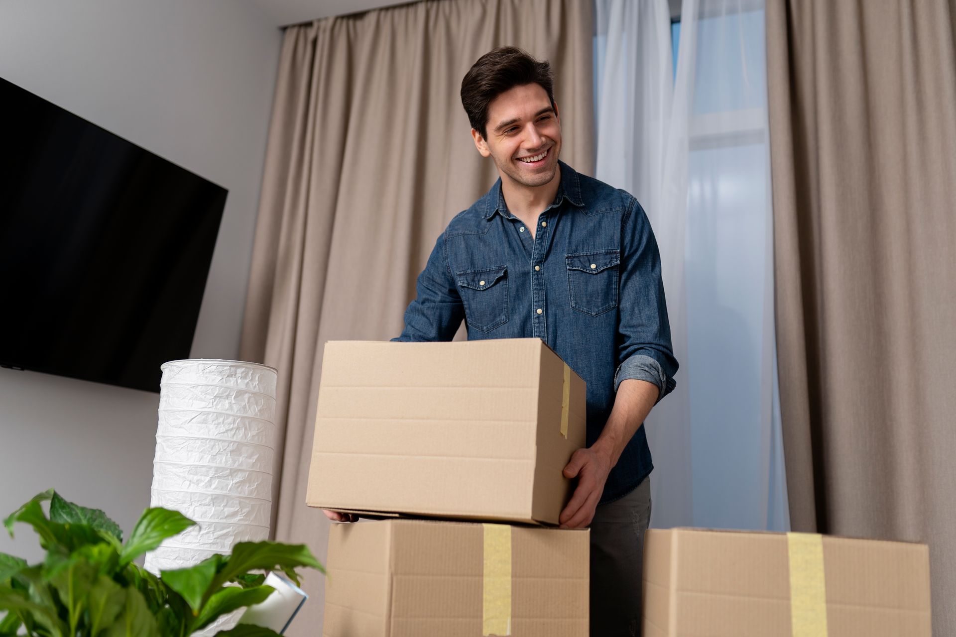 Man smiling, holding cardboard boxes indoors near a TV and window with curtains.