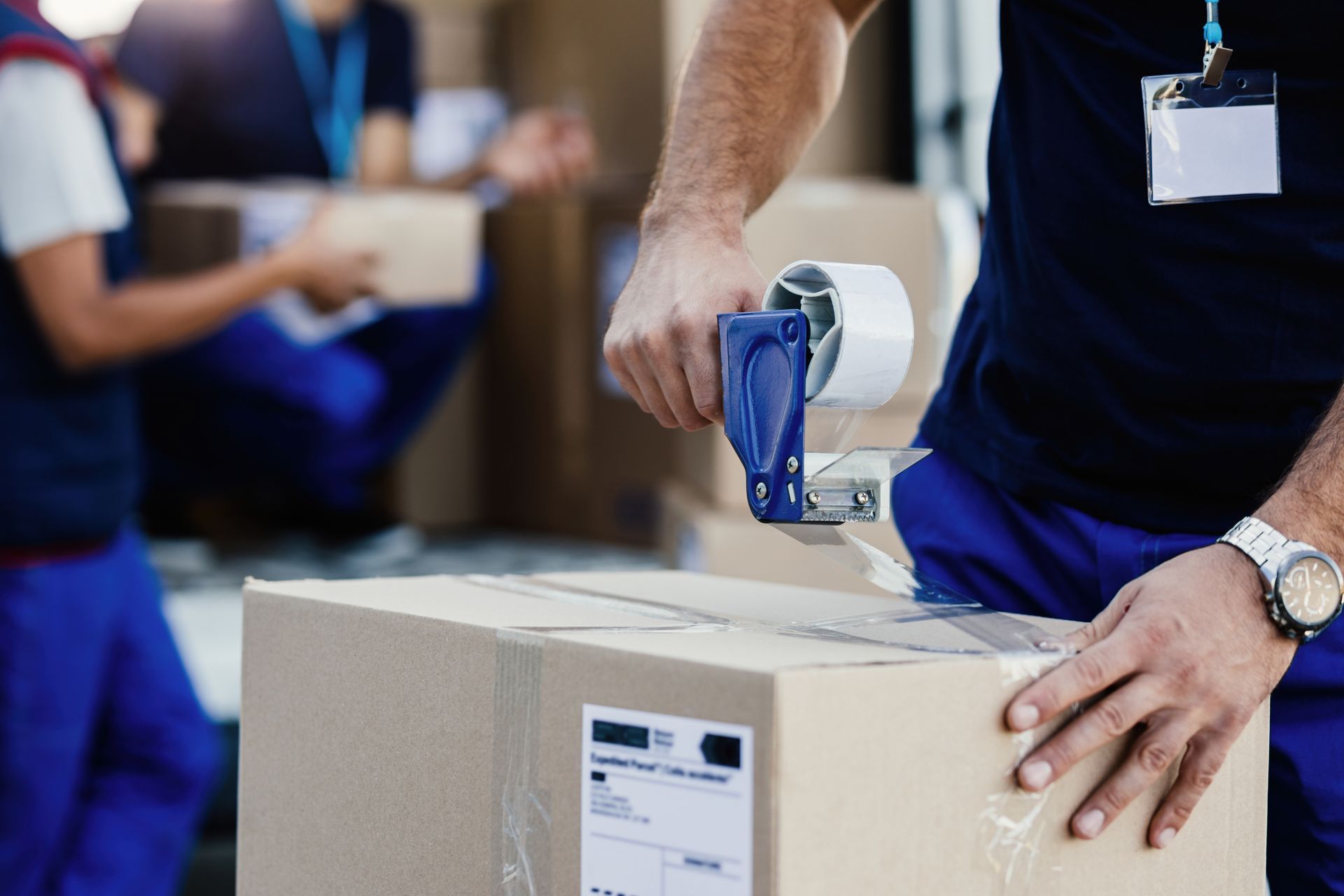 Person seals a cardboard box with tape; others in background pack.