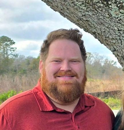 A man with a beard and a red shirt is smiling in front of a tree.
