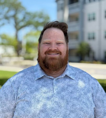 A man with a beard and a red shirt is smiling in front of a tree.