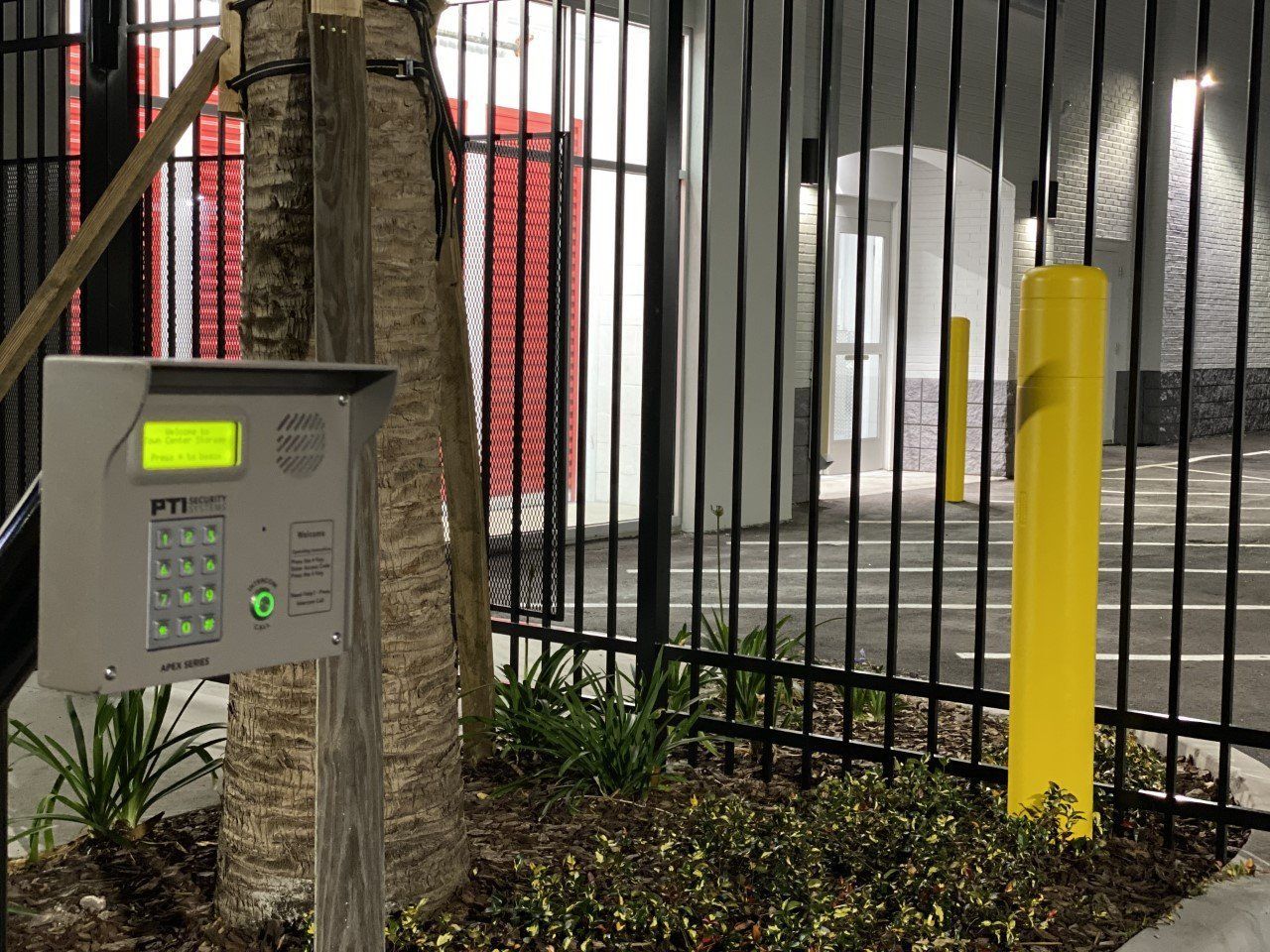 A keypad is sitting next to a yellow pole in front of a fence.