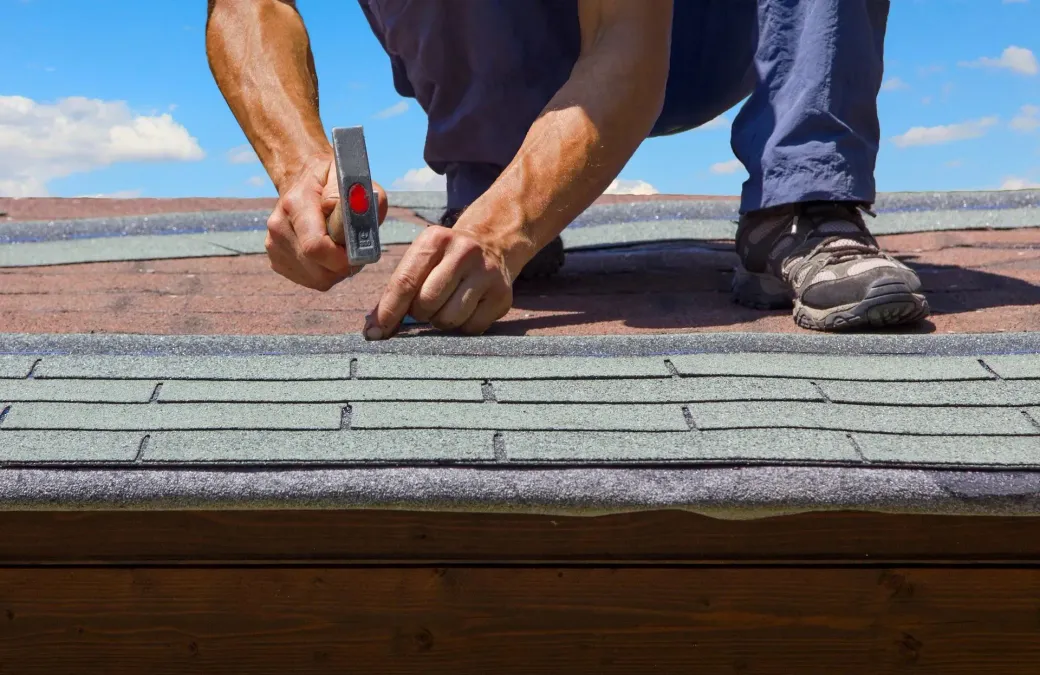 Worker kneeling on a roof, using a hammer to repair gray shingles.