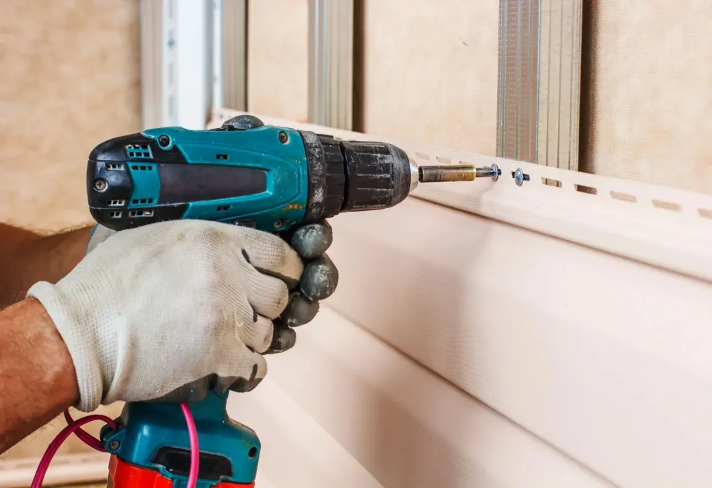 Gloved hands using a teal cordless drill to fasten white siding on a house exterior