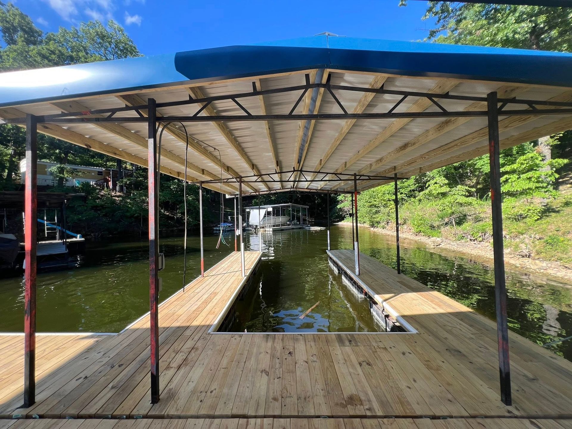 Dock with covered boat slips, wood deck, metal frame, blue canopy, on a lake, surrounded by trees.
