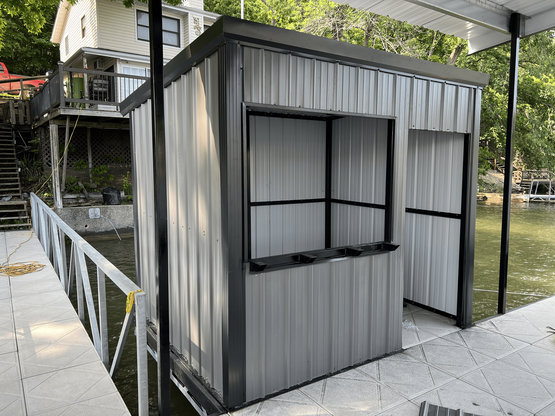 Gray metal shed with a window and open doorway on a dock, next to a lake.