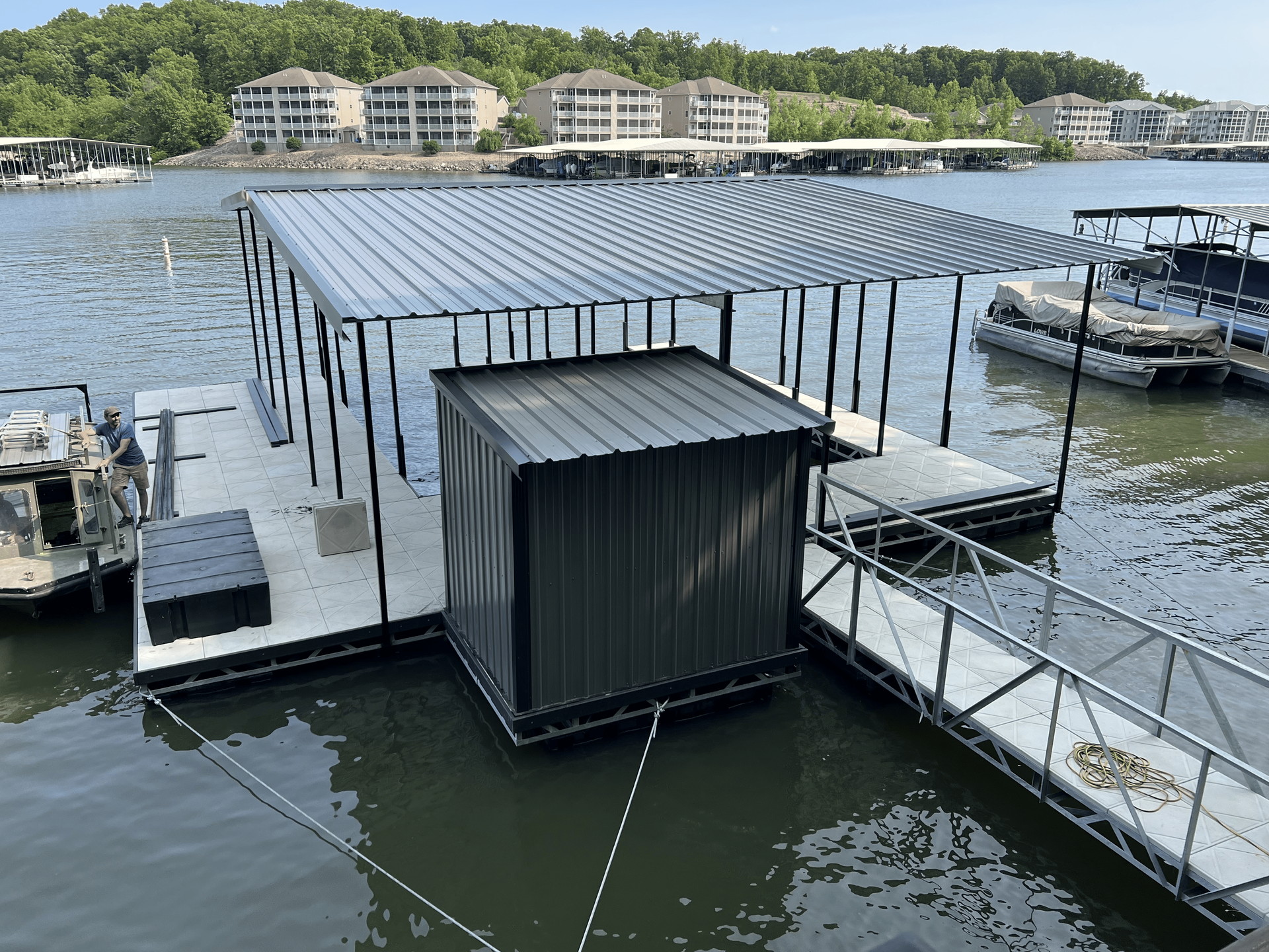 Boat dock with covered slip and metal shed on a lake, residential buildings in the background.