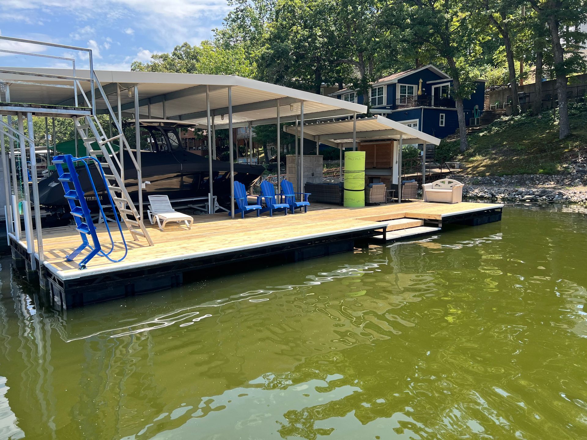 Dock with covered boat slips, blue chairs, and a blue ladder leading into green water.