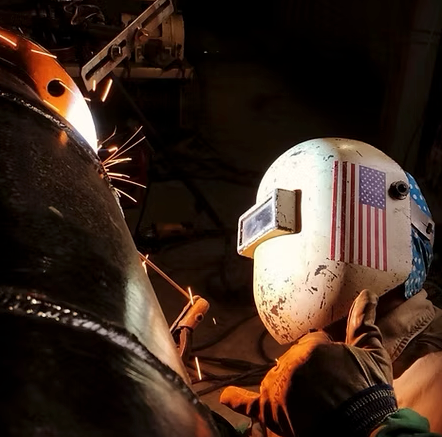 Welder with American flag on helmet working on metal pipe, sparks flying in workshop.
