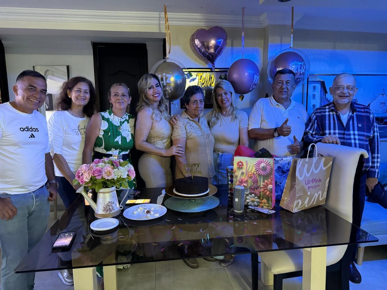 Un grupo de ocho personas se reunió alrededor de una mesa de comedor para una celebración, con pastel y globos de fondo.