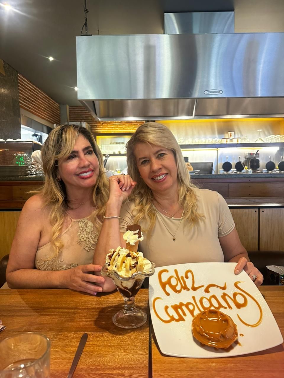 Dos personas sonrientes están sentadas en la mesa de un restaurante con un helado y un plato de cumpleaños escrito con salsa de caramelo.
