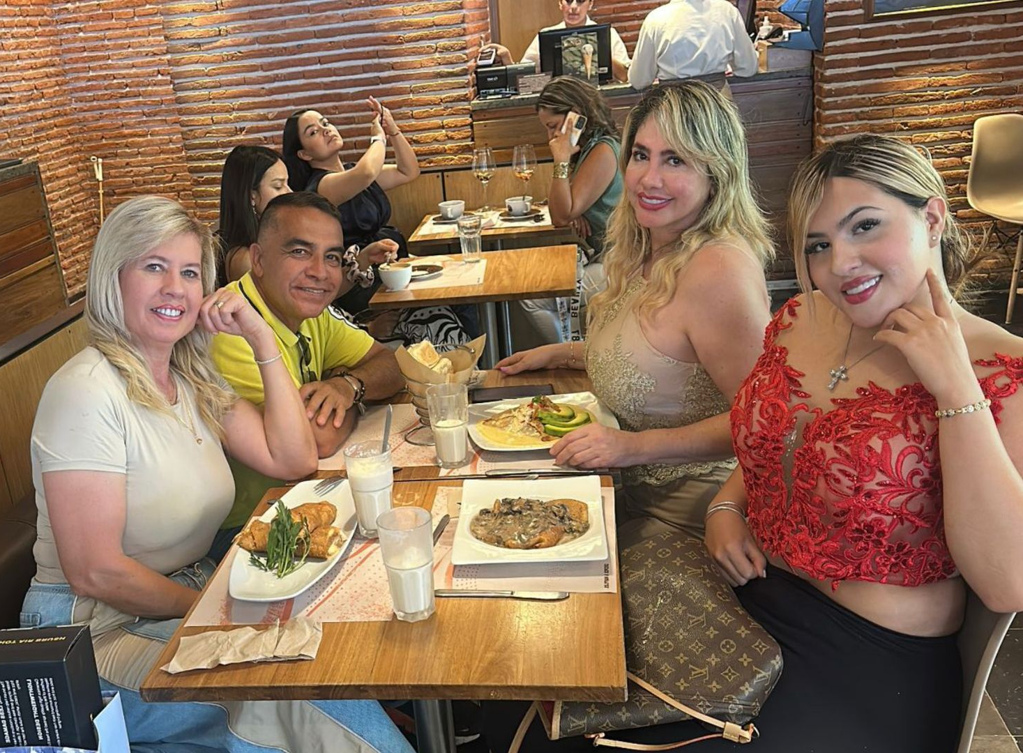 Un grupo de cinco personas sentadas en mesas en un restaurante rústico con paredes de ladrillo, sonriendo y compartiendo una comida.