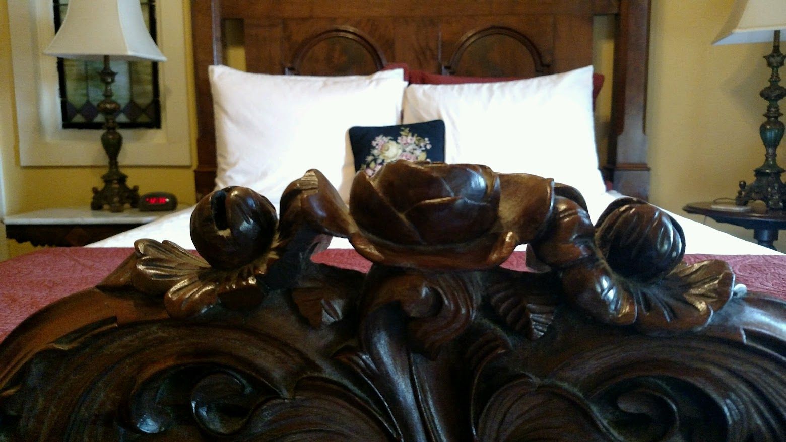 Ornate wooden bed frame with a rose carving, in a hotel room setting with white pillows and lamps.