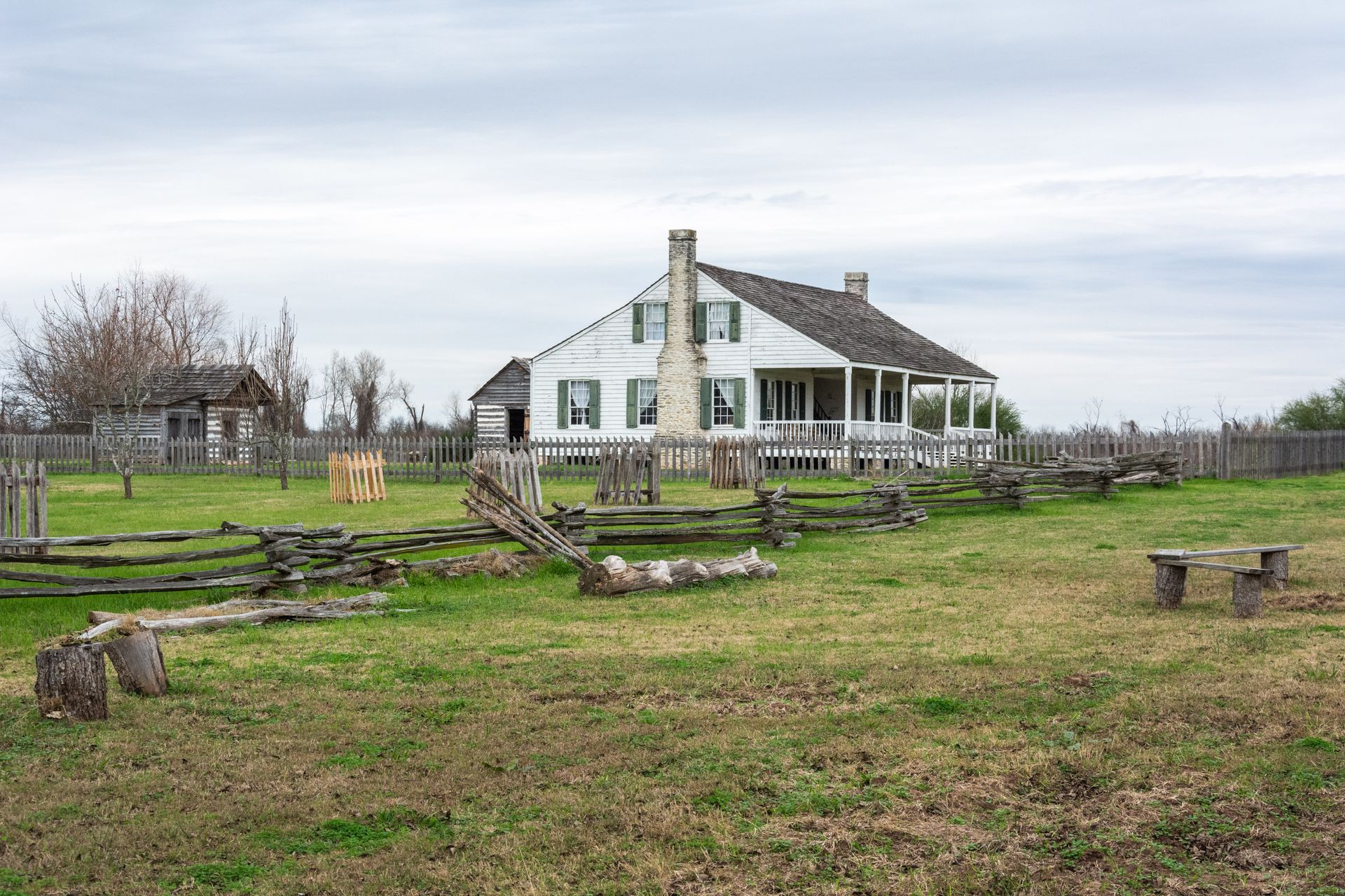 A white, one-story historic house with a stone chimney stands in a grassy field behind a split-rail wooden fence.