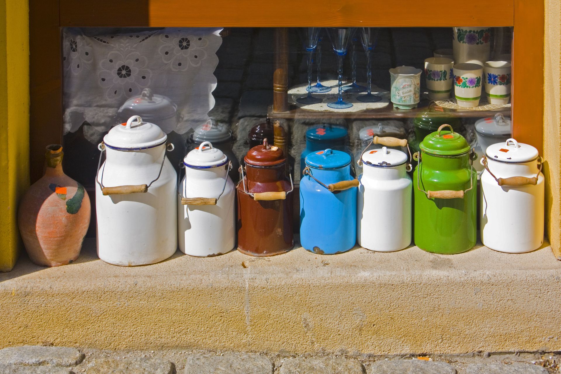 A row of colorful vintage milk cans with wooden handles sit on a window sill, displayed in a shop window.