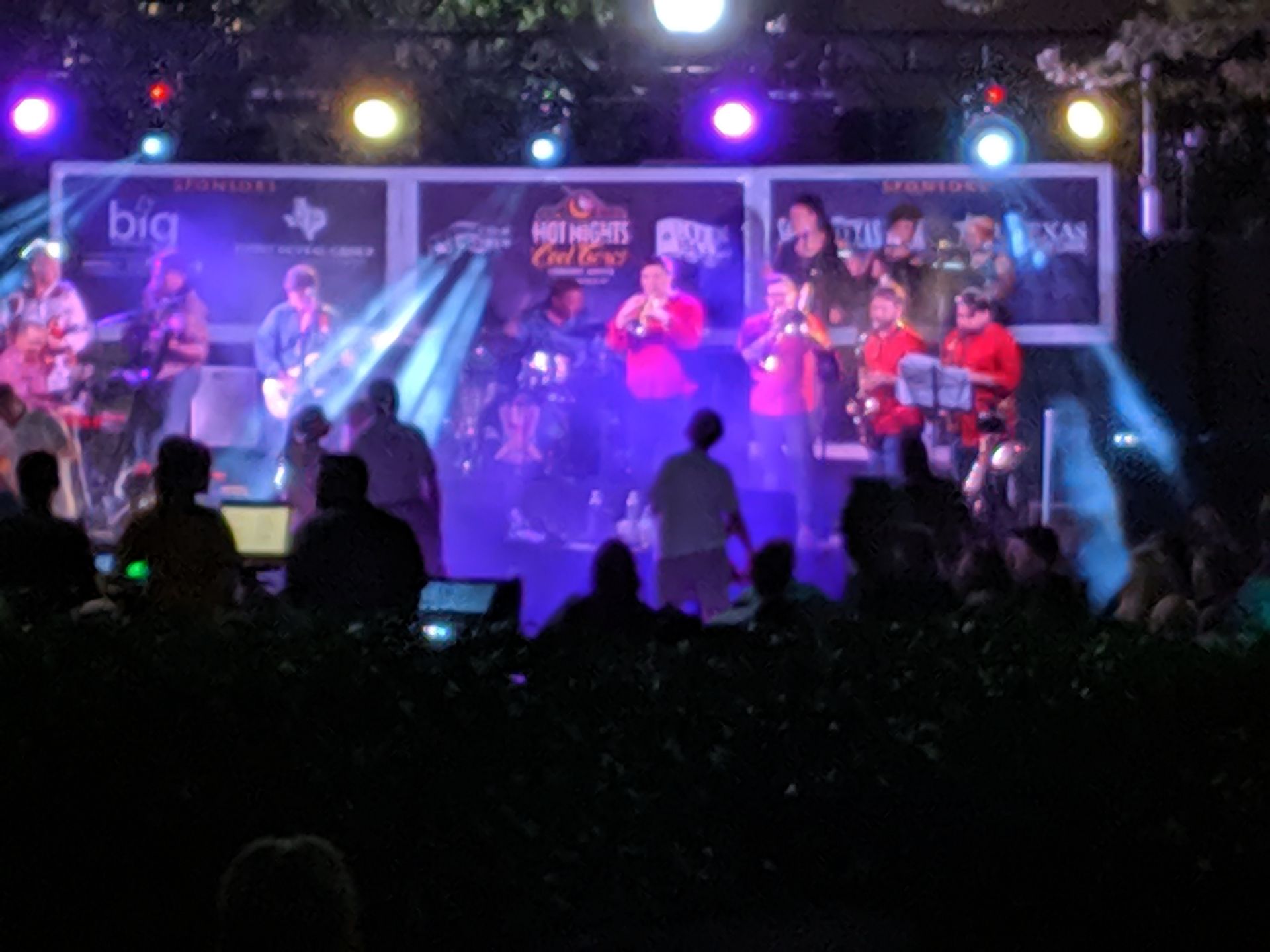 Band performing on a stage at night with spotlights and audience in front.