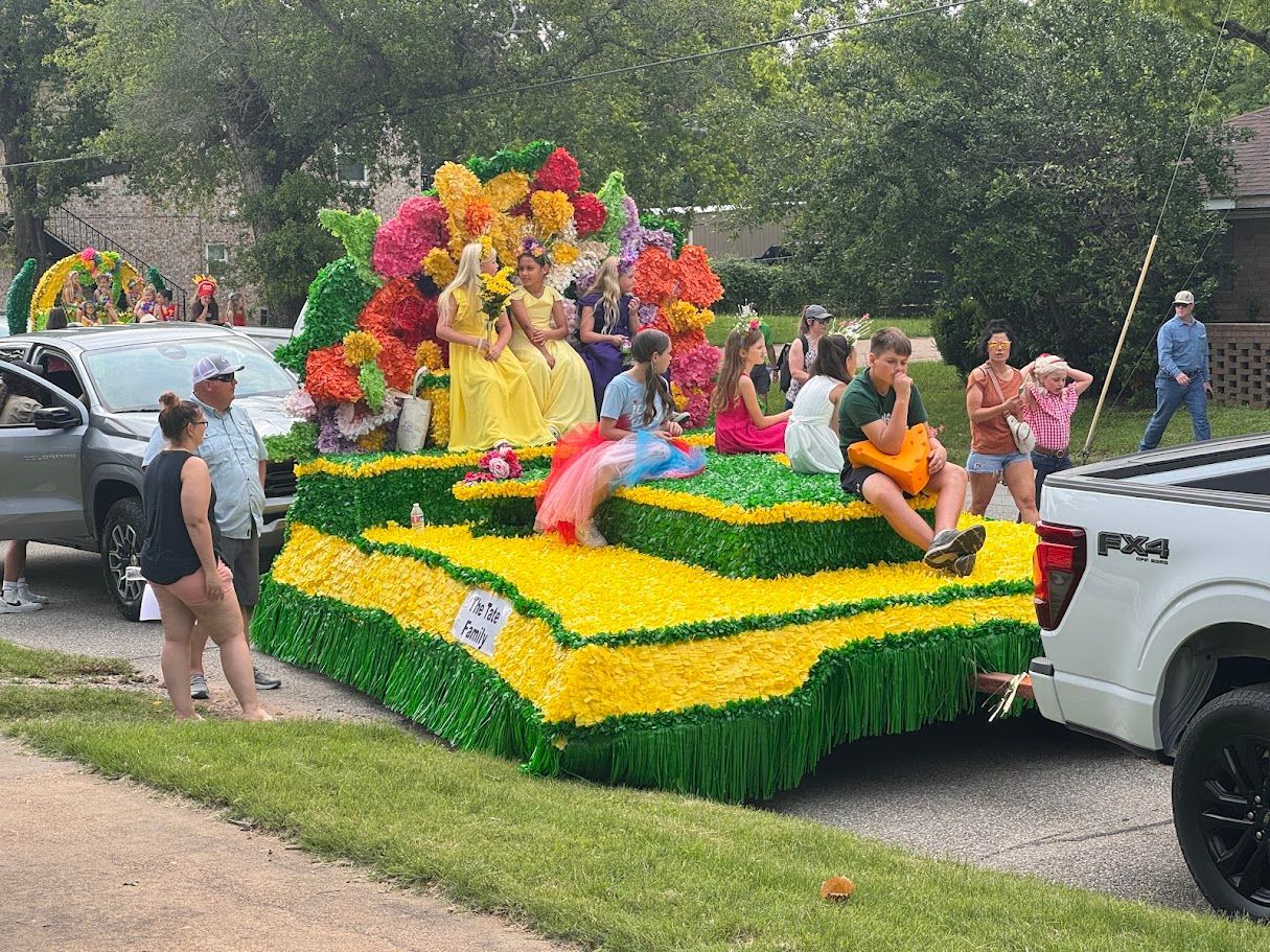 Parade float with people decorated in yellow and green floral arrangements, driving down a street.